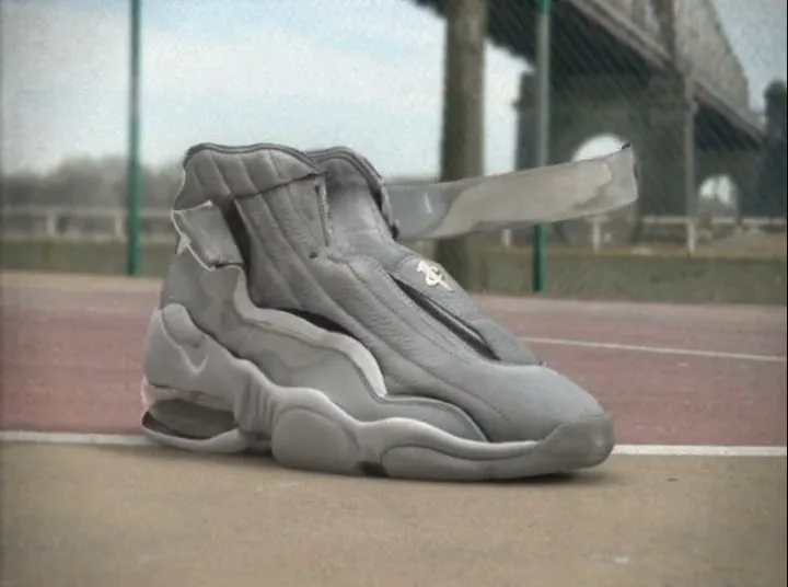 A grey Nike sneaker assembles itself from pieces on a tennis court, with a bridge visible in the background.