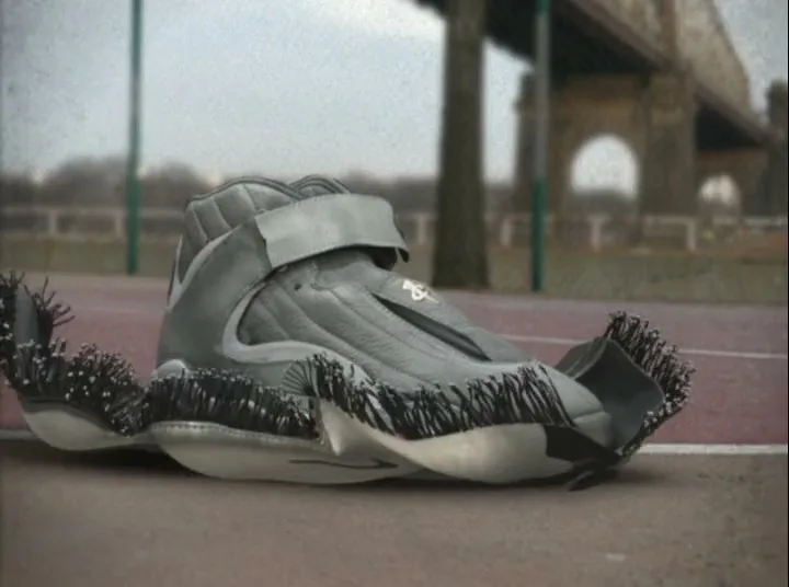 A grey Nike basketball shoe with black bristles on its sole rotates on a red outdoor court, with a bridge in the blurry background.