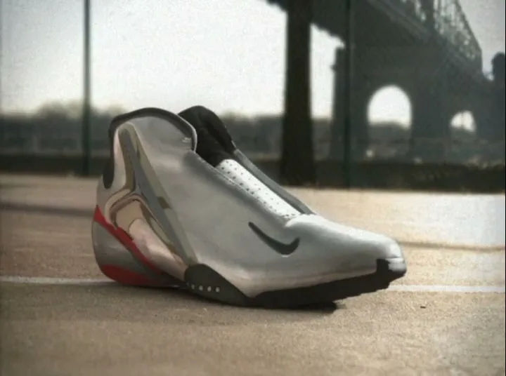 A silver and black Nike basketball shoe with red accents sits on a concrete surface, with a bridge in the background.