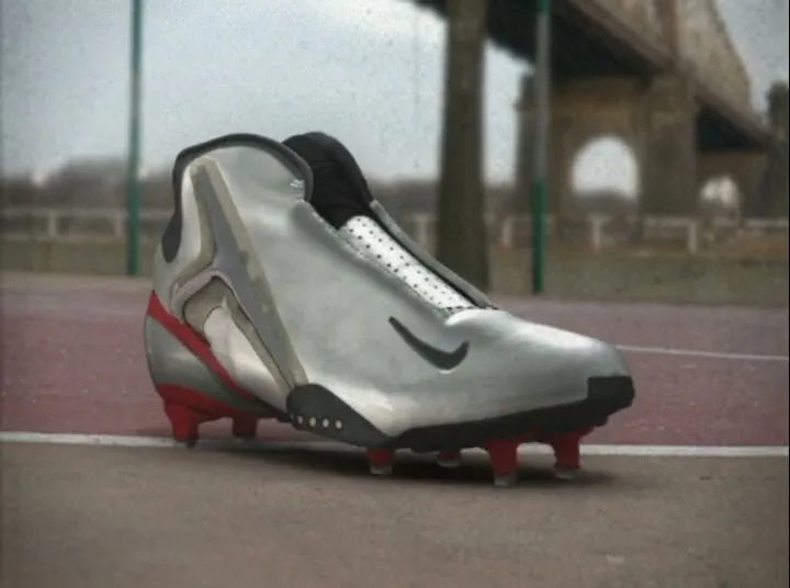 A silver and red Nike football boot with studs is shown on a red and grey outdoor court, with a bridge in the background.