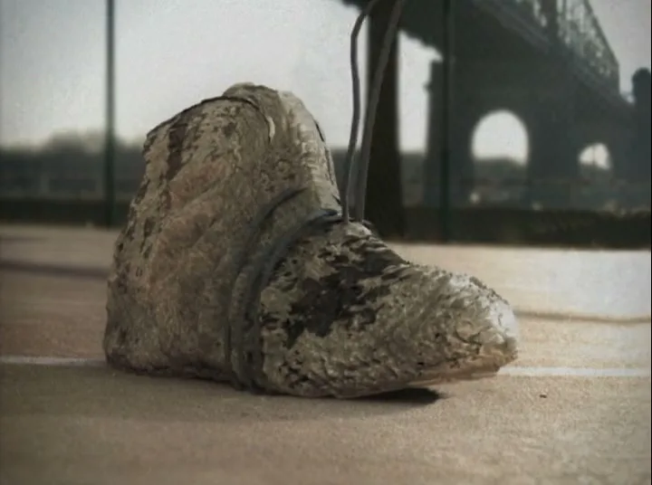 A stone shoe transforms into a modern athletic shoe with laces wrapping around it, on a concrete surface with a bridge in the background.
