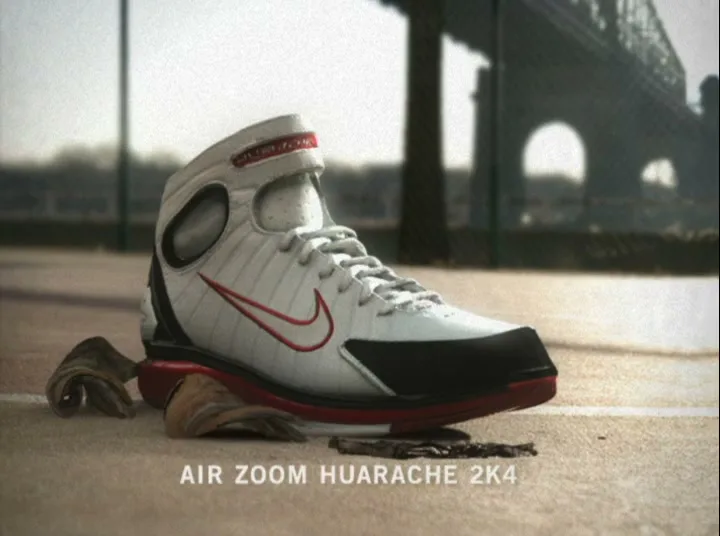 A white and red Nike basketball shoe assembles itself from pieces on a concrete court, with a bridge in the blurry background.