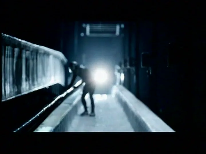A lone figure in a dark bobsled track bends down, illuminated by a bright light in the distance, creating a dramatic silhouette.