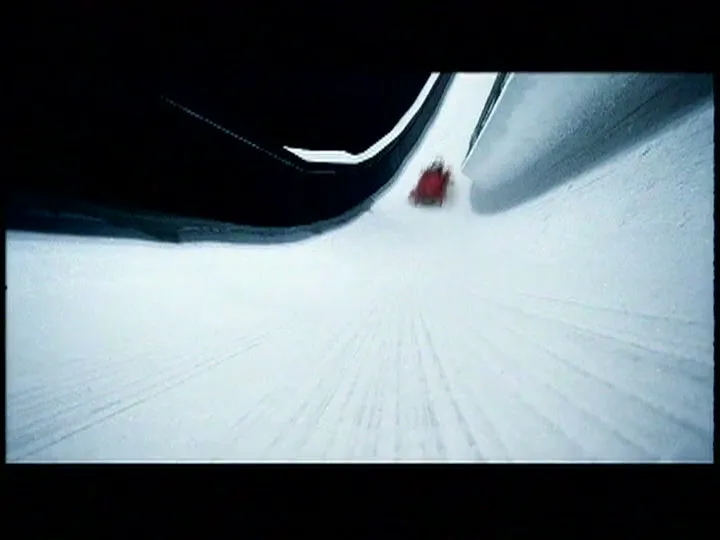 A red bobsled with two people speeds down an icy track, seen from a low angle as it approaches the camera.