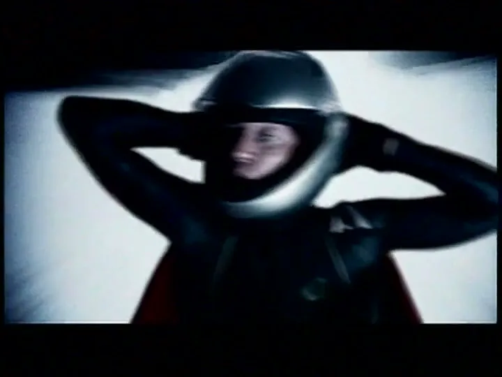 A male bobsledder in a black suit and silver helmet adjusts his helmet with both hands, looking intense in a bobsled.