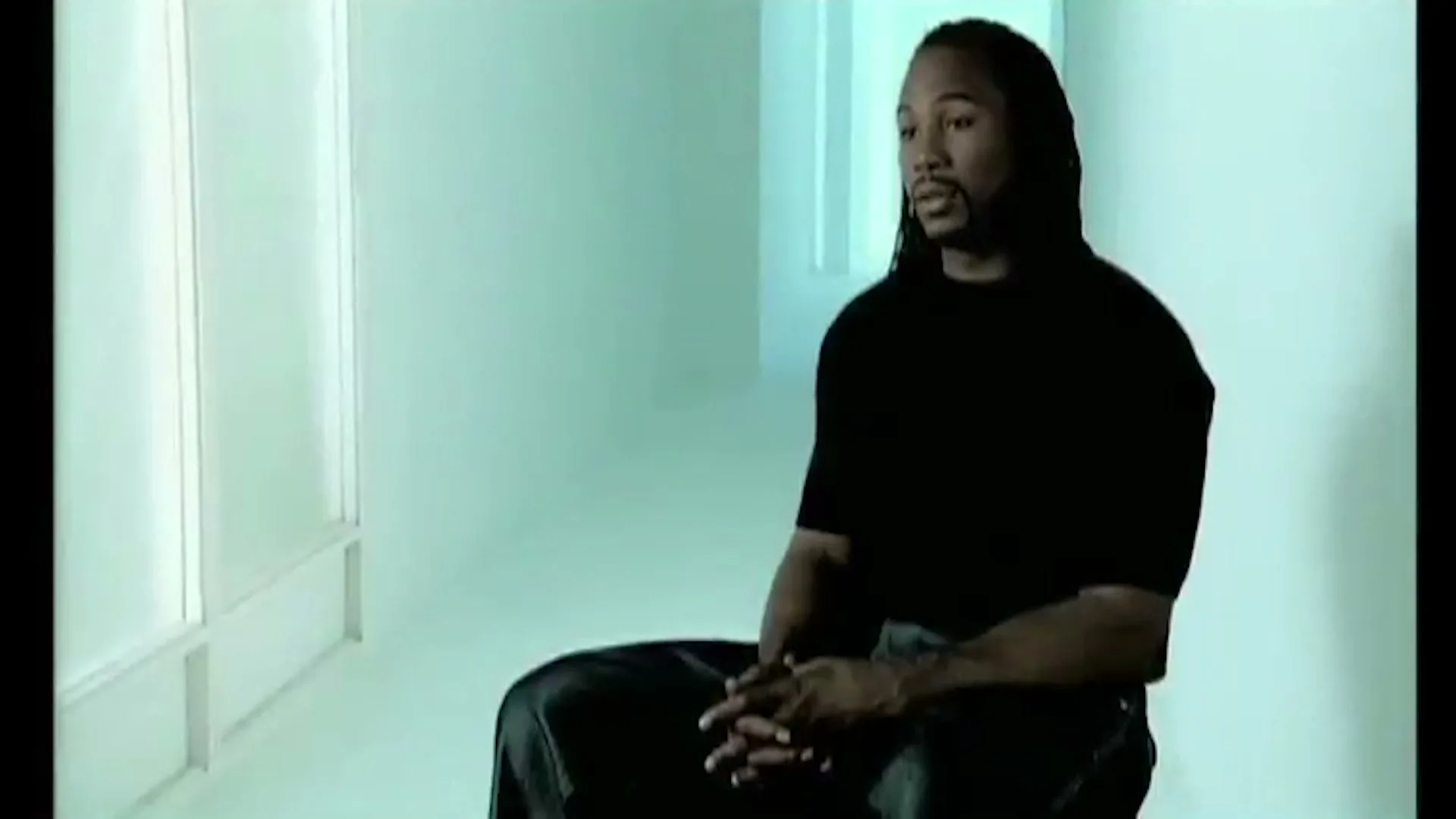 A man with long dreadlocks, wearing a black t-shirt, sits and speaks in a brightly lit, minimalist room.