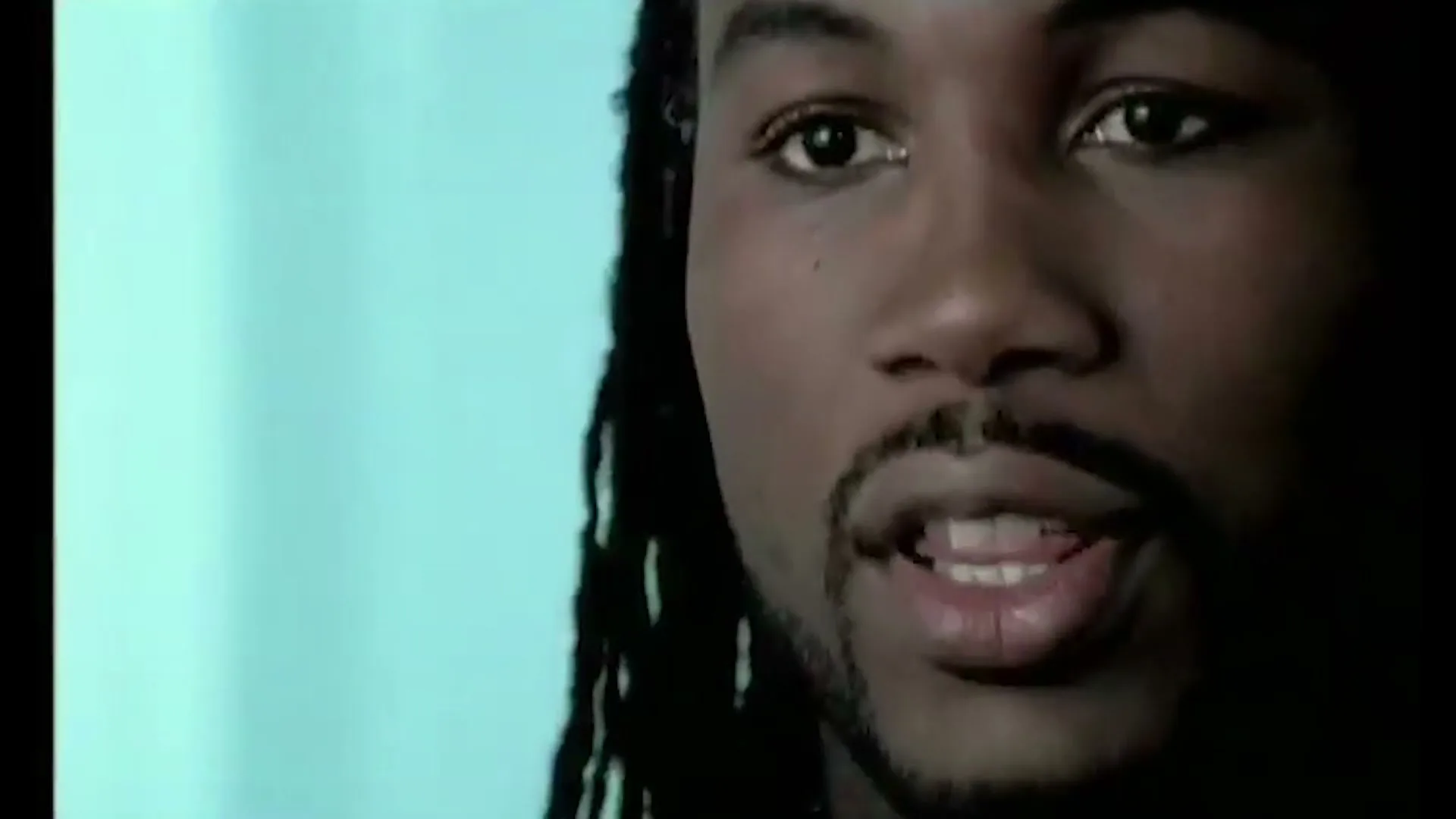 A man with dreadlocks and a goatee speaks directly to the camera in a medium closeup shot, with a faint overlay of another person's face.