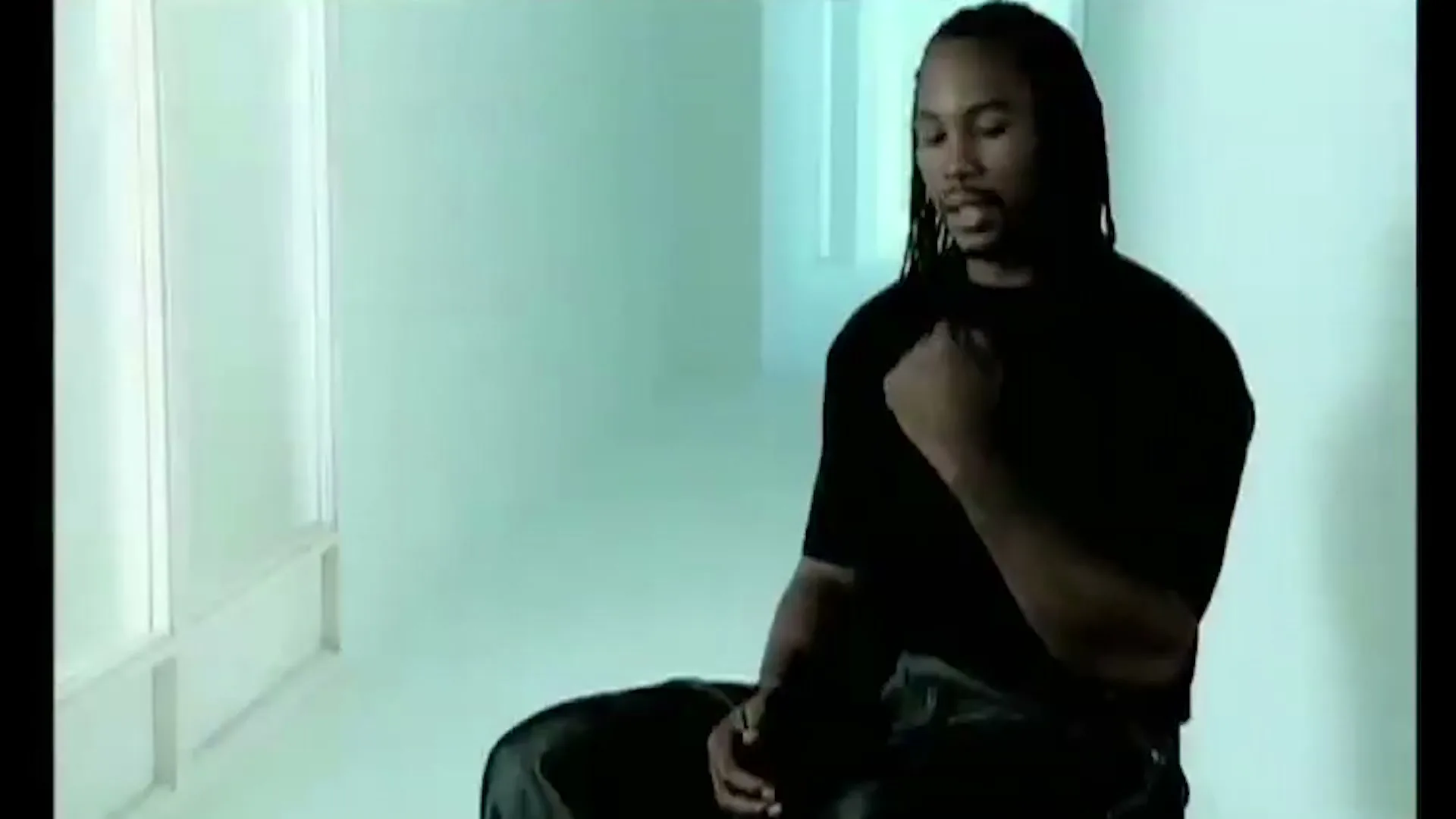 A man with dreadlocks, wearing a black t-shirt, sits and looks to the right in a bright, minimalist white room.