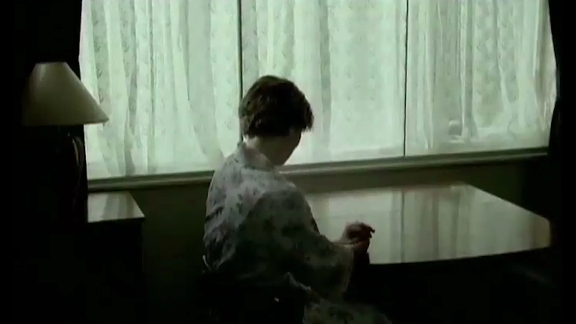A woman in a floral robe sits with her back to the camera, looking out a window with sheer curtains in a dimly lit room.