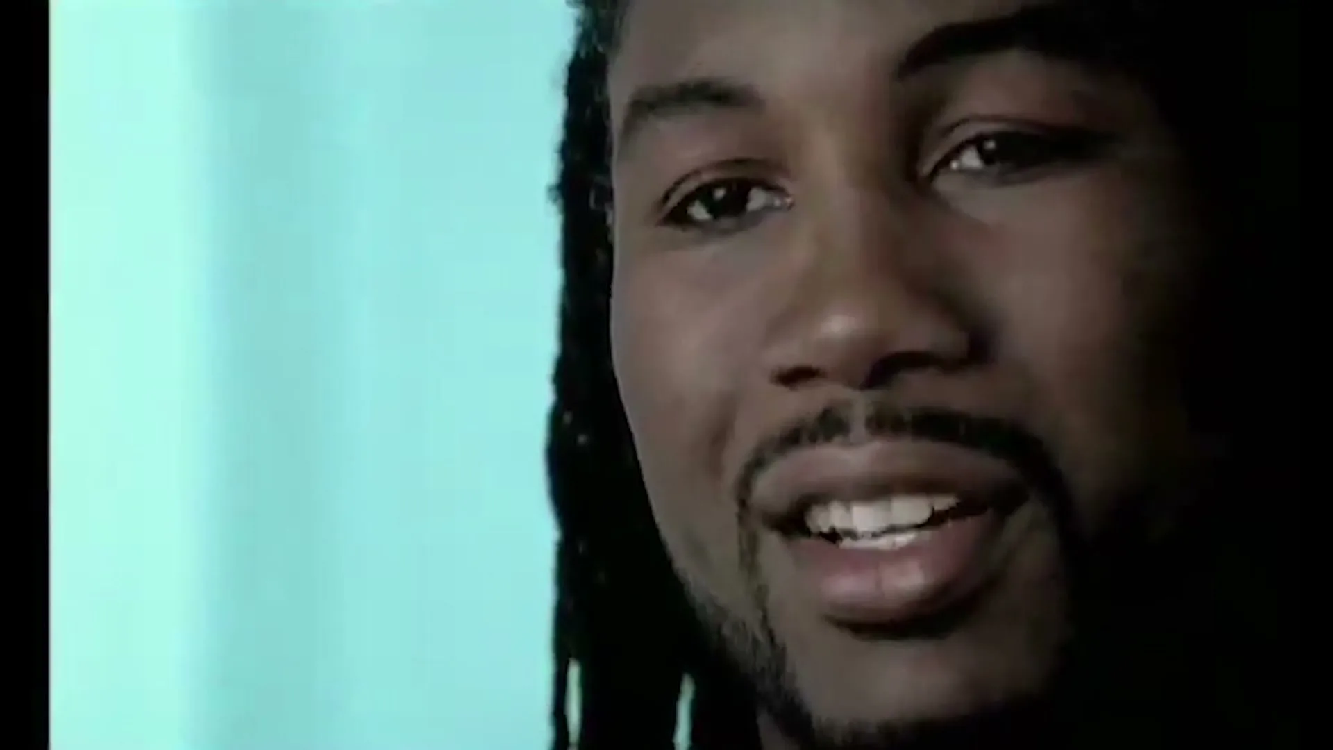 A man with dreadlocks and a goatee speaks to the camera in a medium closeup shot, with a light blue background.