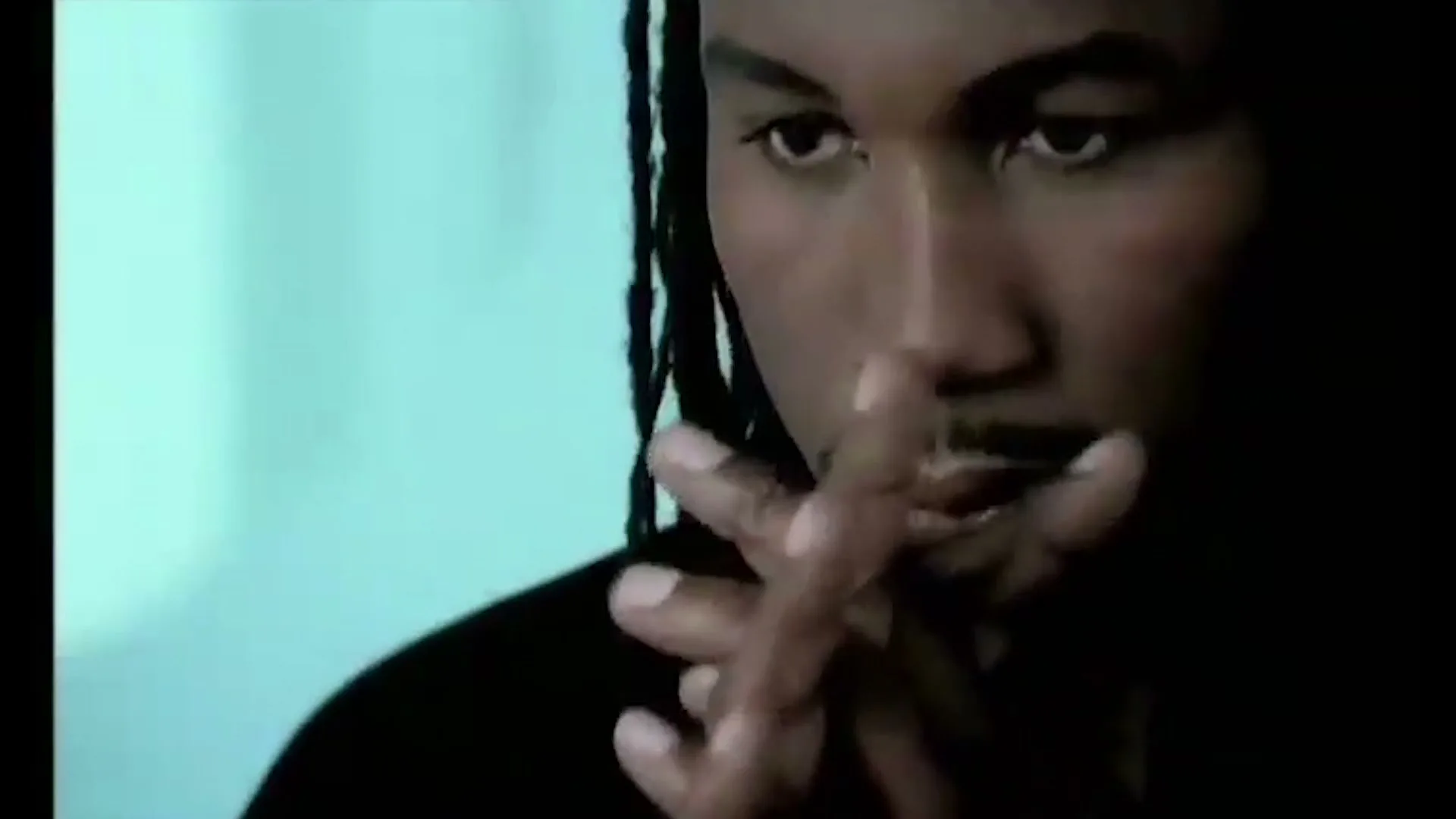 A man with dreadlocks and a mustache looks down, with his hands clasped in front of his mouth, in an interior setting.