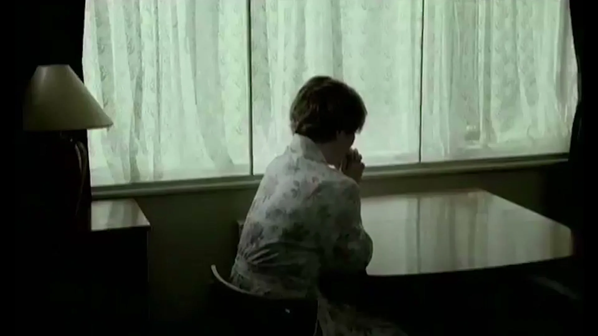 A woman in a floral robe sits at a table, looking down with her hands clasped, in a dimly lit room with a window.