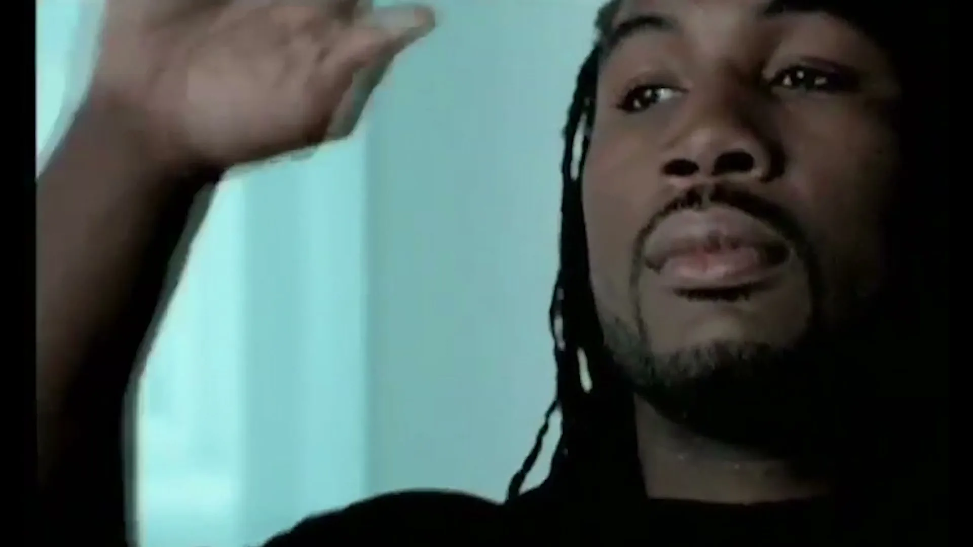 A Black man with dreadlocks and a goatee looks up and raises his hand to his head in an interior setting.