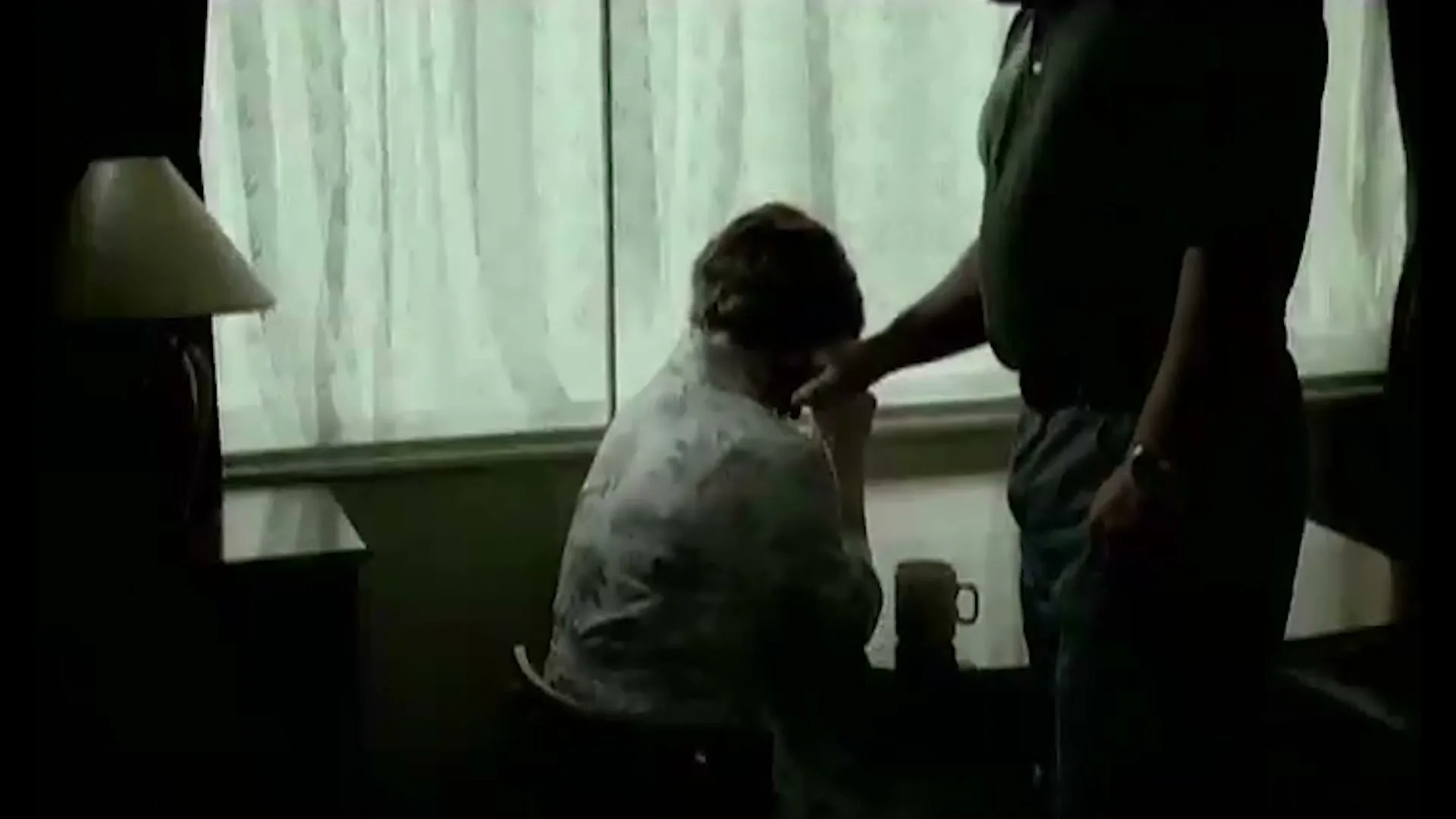 A man in a dark polo shirt comforts a woman in a floral shirt who is sitting and crying by a window in a dimly lit room.