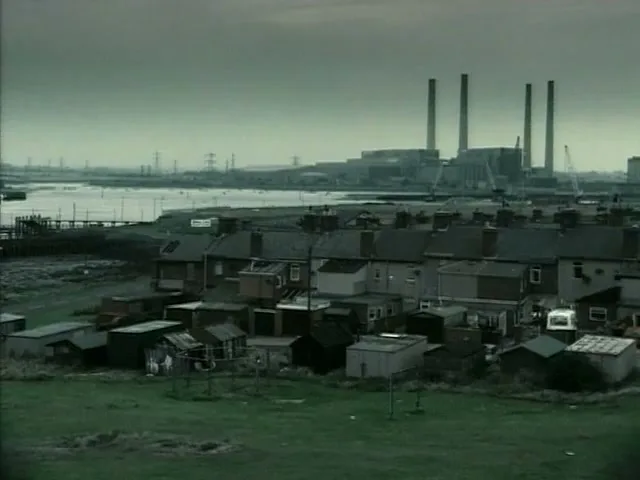 A wide shot of a rundown coastal town with small houses and sheds, with a large power plant and industrial area in the background under a grey sky.