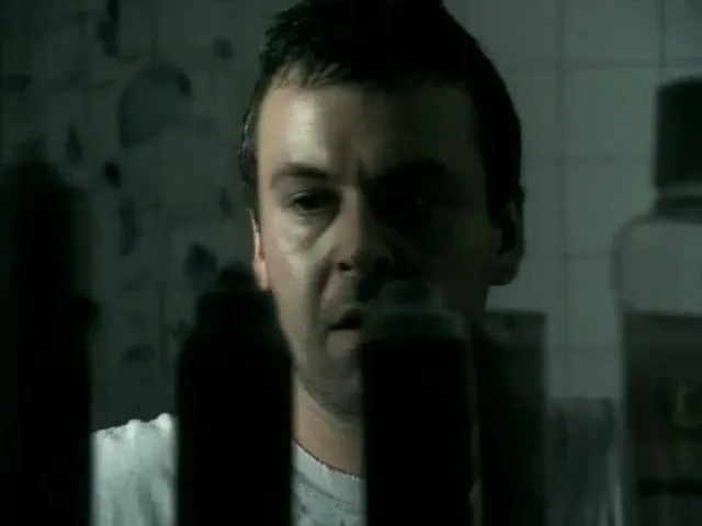 A man with dark hair and a white t-shirt looks shocked, reflected in a mirror with bottles in the foreground in a bathroom.