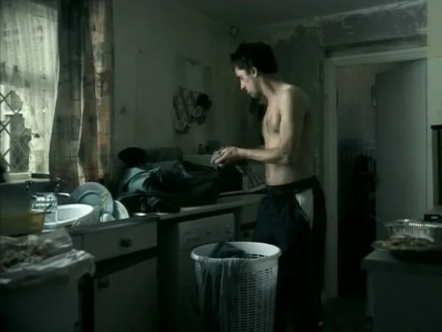 A shirtless man in his 20s stands in a messy, run-down kitchen, putting clothes into a laundry basket.