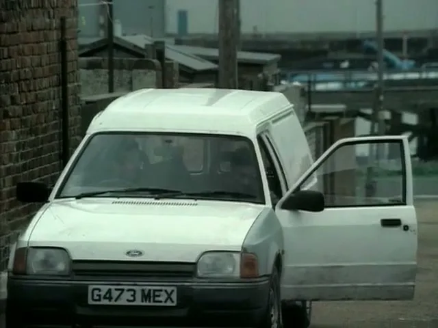 A man in a dark jacket and pants gets into a white Ford van parked on a narrow street next to a brick wall.