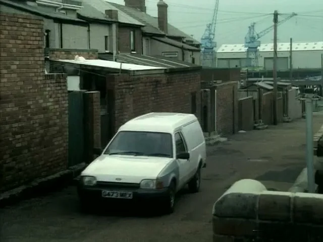 A white Ford van drives slowly down a narrow, brick-walled alleyway in an industrial area with houses and cranes visible in the background.