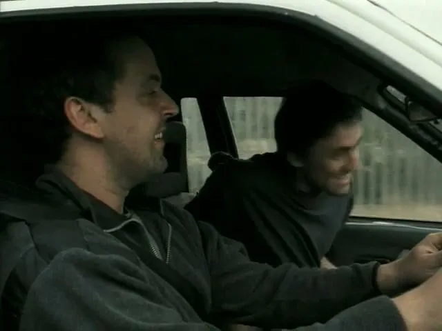 Two men in a car, the driver smiling and the passenger looking out the window, driving on a road during the day.