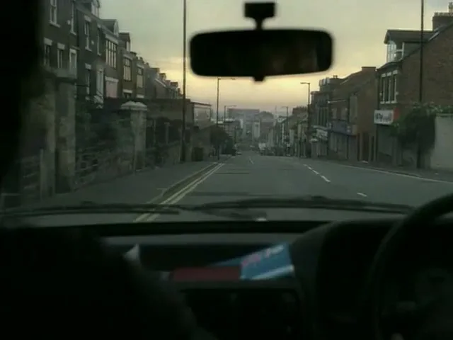 A POV shot from inside a car driving down a street lined with houses and shops, with a rearview mirror visible.