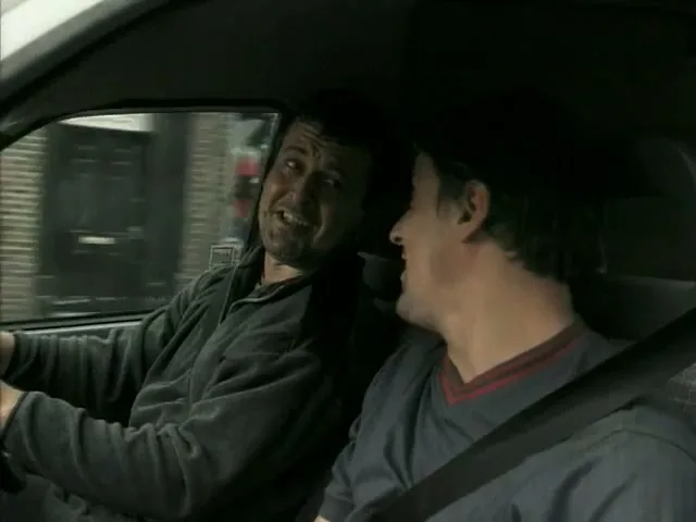 Two men are driving in a car, the driver looks worried while the passenger sings loudly, as they drive past buildings.