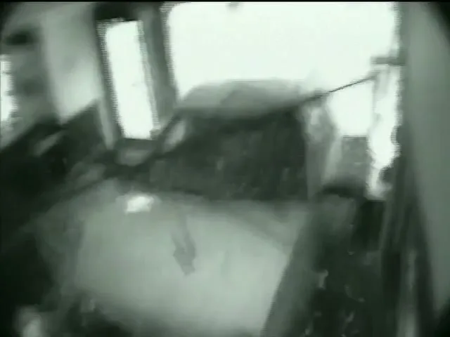 A blurry, grainy, black and white security camera shot shows a car crashing through a storefront window, sending debris flying into the interior.