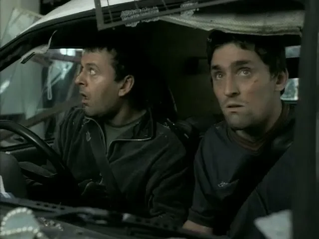 Two men in a wrecked car look out the windshield with wide eyes, appearing shocked and scared.