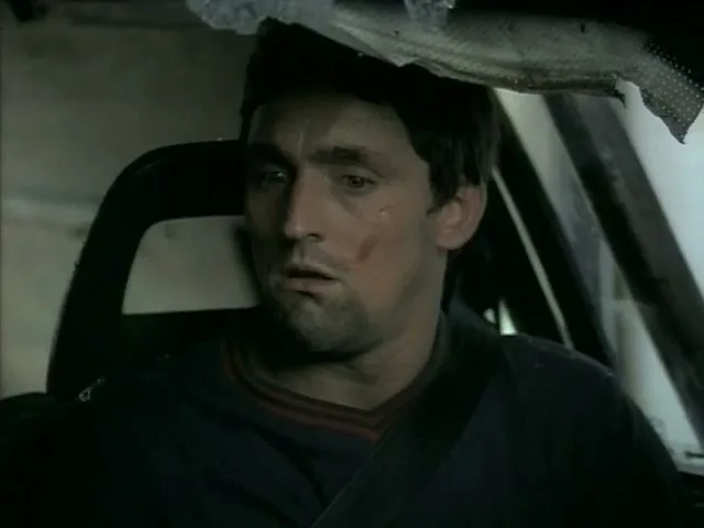 A man with a bruised face and dark hair sits in a car, looking around with a worried expression after a car crash.