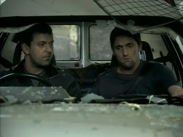 Two men, one driving and one in the passenger seat, look at each other with worried expressions inside a damaged vehicle.