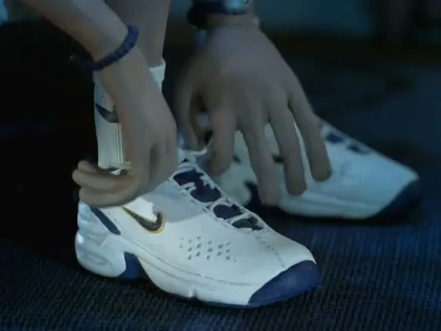 A young person with light skin and a blue bracelet ties the laces of a white and blue Nike sneaker on a dark floor.