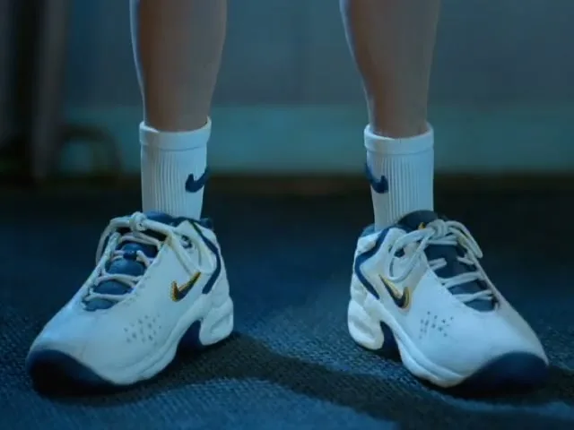 A child's legs and feet are shown as they put on white and blue Nike sneakers with white socks, on a dark floor.