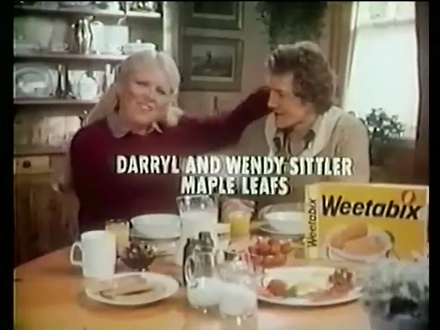 A blonde woman in a maroon sweater puts her arm around a man in a beige shirt at a breakfast table with a box of Weetabix.
