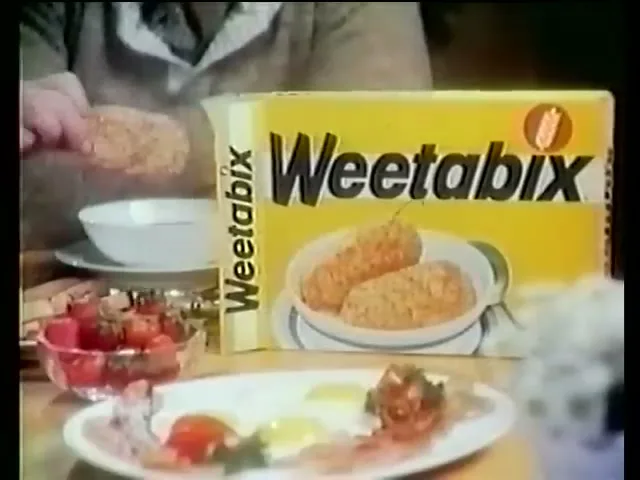 A person's hands are seen placing a Weetabix biscuit into a bowl, with a box of Weetabix and a plate of breakfast food on a table.