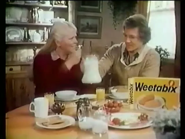A woman with blonde hair and a man with curly brown hair sit at a breakfast table, as the man pours milk into a bowl of cereal.