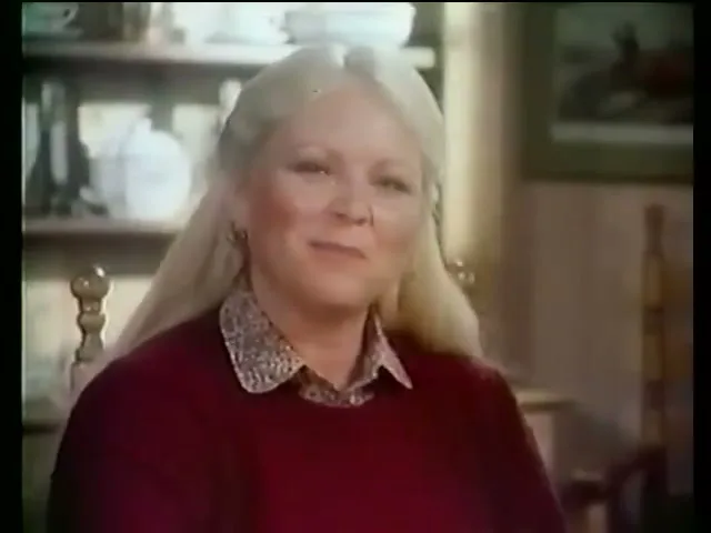 A blonde woman in a red sweater and patterned collar smiles and looks off-camera, holding a small fruit and a spoon in a cozy interior setting.