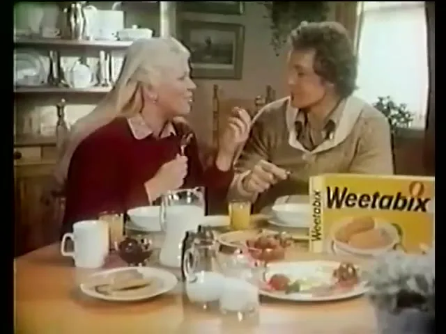 A blonde woman and a man with curly hair are seated at a breakfast table, talking and eating cereal, with a box of Weetabix visible on the table.