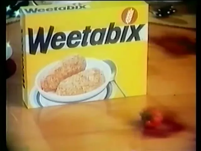 A box of Weetabix cereal sits on a wooden table as a strawberry rolls into frame and stops next to it.