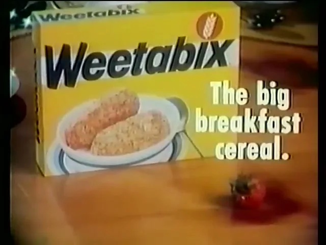 A box of Weetabix cereal is displayed on a wooden table with two cereal biscuits in a bowl and a strawberry, with text "The big breakfast cereal."