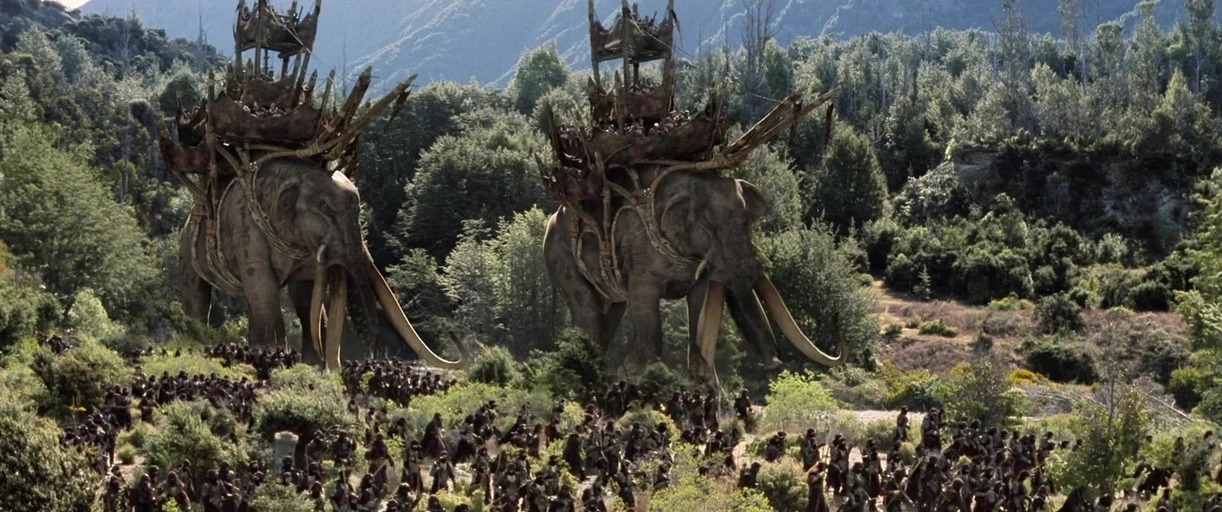 A dense, mountainous valley filled with green shrubs and trees during the day. An army of armored soldiers walks alongside two giant, tusked war elephants.