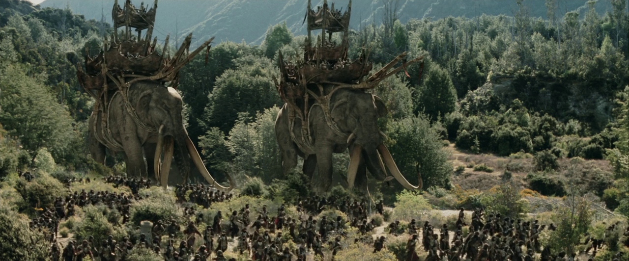 A dense army of soldiers marches through a verdant, mountainous valley floor. Two giant war elephants equipped with wooden platforms traverse the landscape among the crowd.