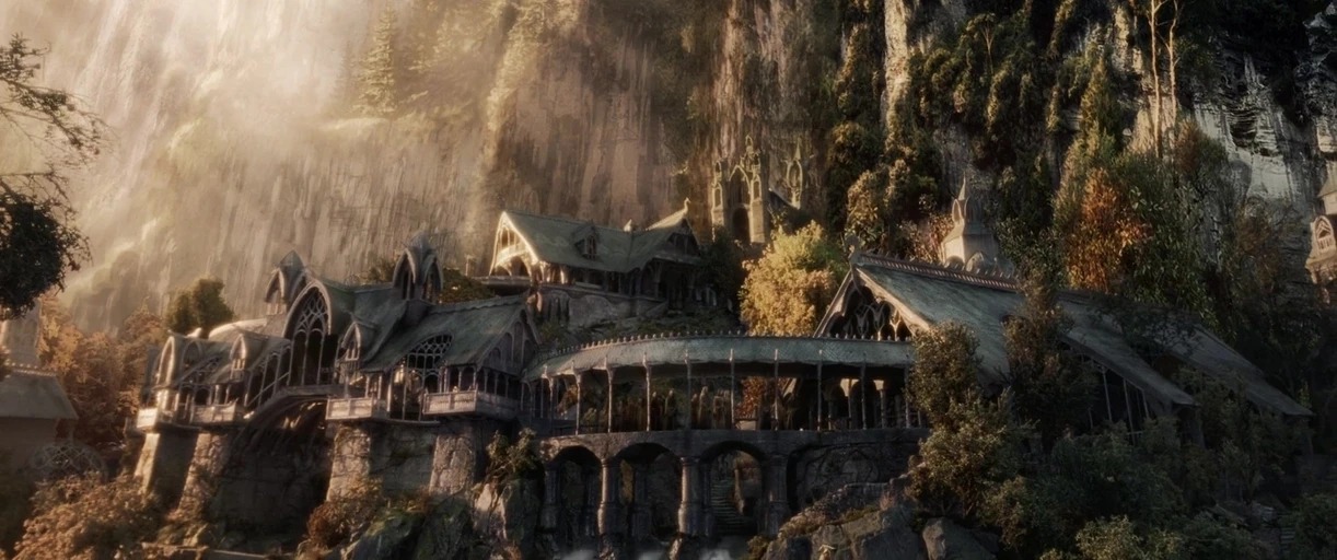 Intricate stone structures with arched roofs sit nestled against a towering forested cliffside. A curved stone bridge connects the buildings over a rocky, flowing stream.