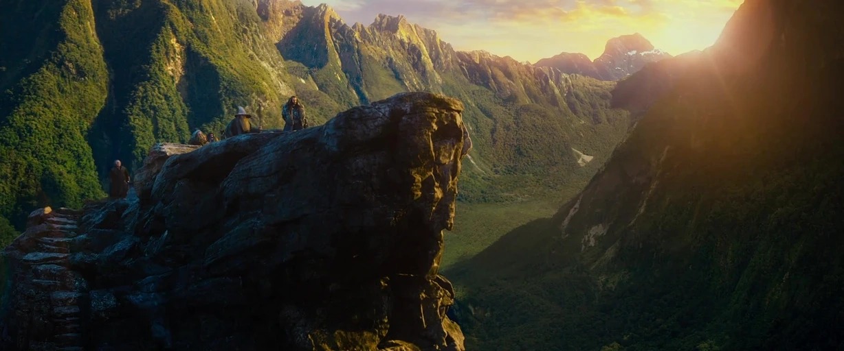 A vast mountain valley features steep slopes covered in dense green vegetation at sunset. A group of people stands on a narrow, exposed rocky ledge in the foreground.