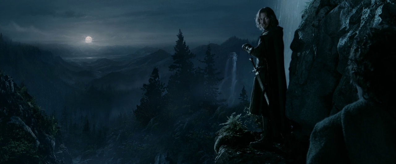 A mountain cliff at night overlooks a vast forested valley under a full moon. A man in a dark cloak stands on the ledge while another figure is visible.