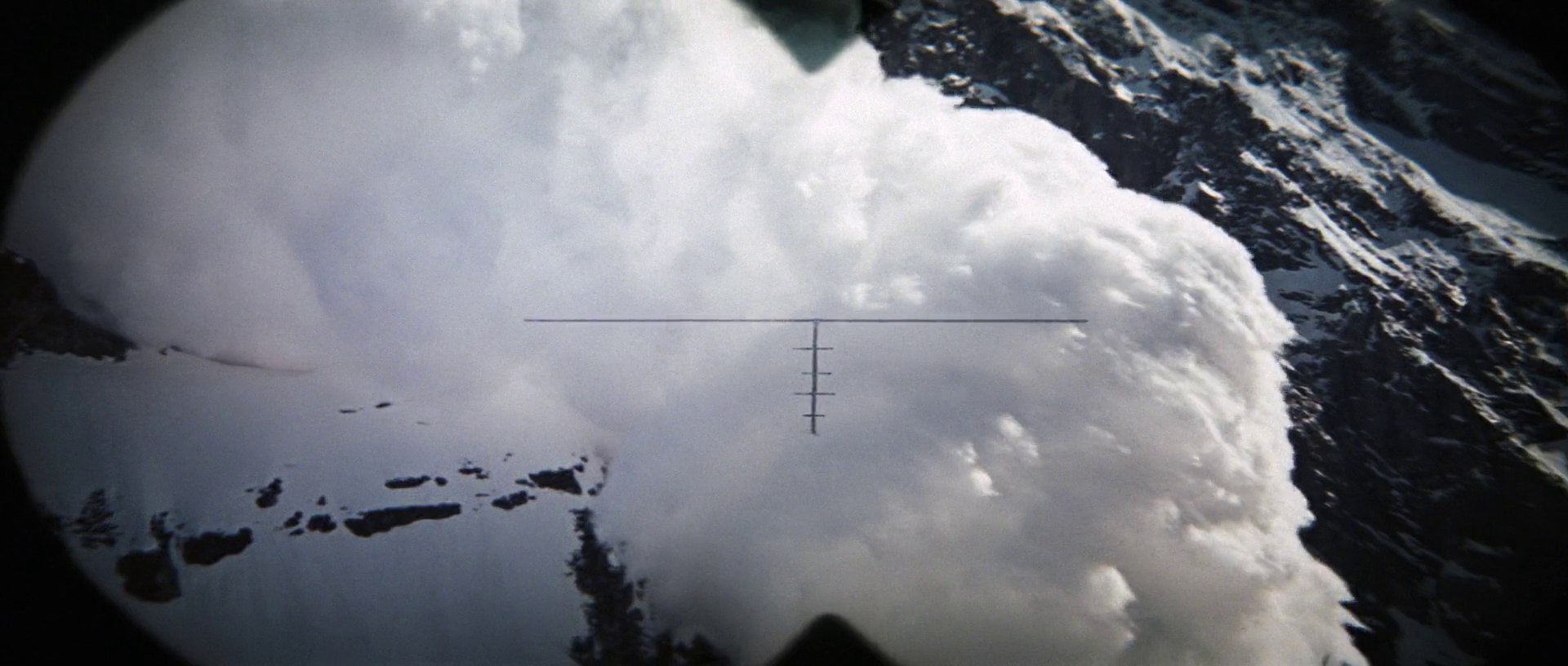 A massive white snow avalanche tumbles down a steep, rocky mountain cliff side. The view is framed through a circular reticle indicating an optical scope.