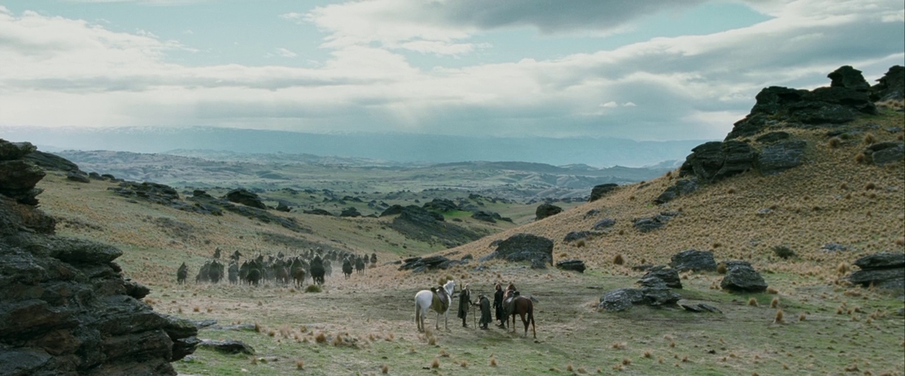 Wide landscape of rolling grassy hills interspersed with jagged black rock formations under overcast. A small group stands with horses while a larger mounted party approaches from the distance.