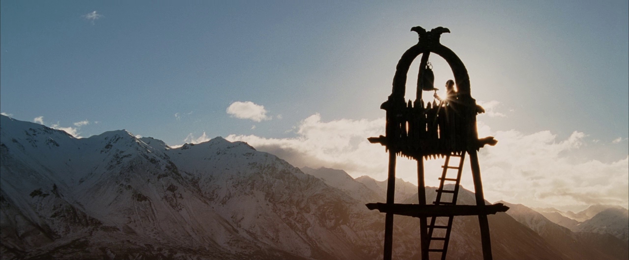 A high, wooden watchtower stands against a backdrop of massive, snow-capped mountain peaks. A lone figure reaches out to strike a bell hanging within the tower structure.