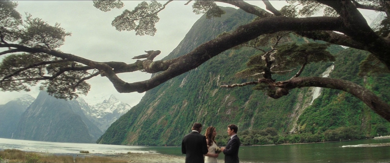A man and woman stand facing each other before a man in a suit. They are on the shore of a vast lake framed by dramatic steep mountain slopes.