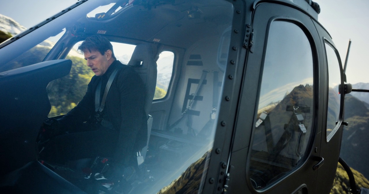 The interior of a helicopter cockpit shows metal framing and grey upholstered seating. A pilot wearing a dark jacket operates the controls while flying over mountainous terrain.