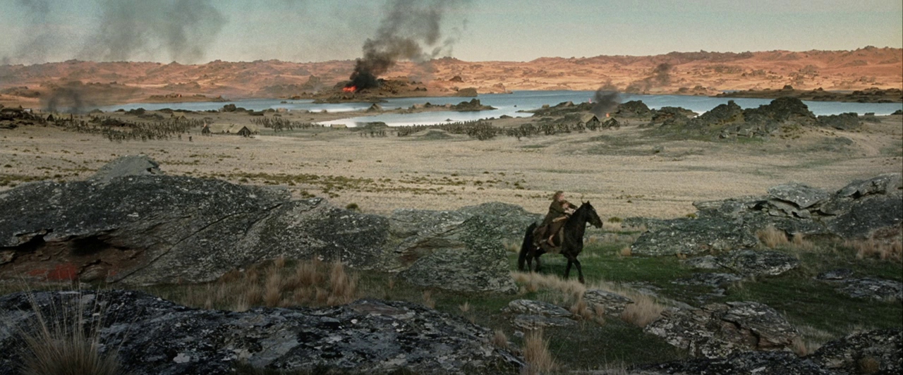 A vast rocky terrain overlooks a lake with burning structures and black smoke plumes. A lone figure on horseback traverses the foreground landscape during a military engagement.